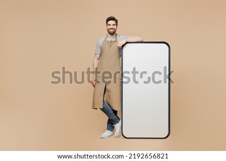 Full body young man barista barman employee wear brown apron work in coffee shop big huge blank screen mobile cell phone isolated on plain pastel light beige background. Small business startup concept Royalty-Free Stock Photo #2192656821