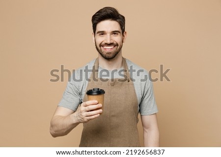 Young cheerful cool man barista barman employee wear brown apron work in shop hold craft paper brown cup coffee to go isolated on plain pastel light beige background. Small business startup concept Royalty-Free Stock Photo #2192656837