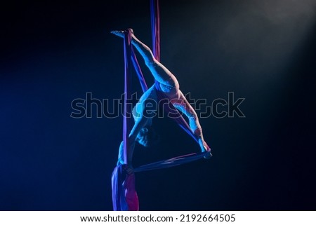 Female circus gymnast hanging upside down on aerial silk and demonstrates stretching. Young woman performs tricks at height on silk fabric. Acrobatic stunts on black background with blue backlight. Royalty-Free Stock Photo #2192664505