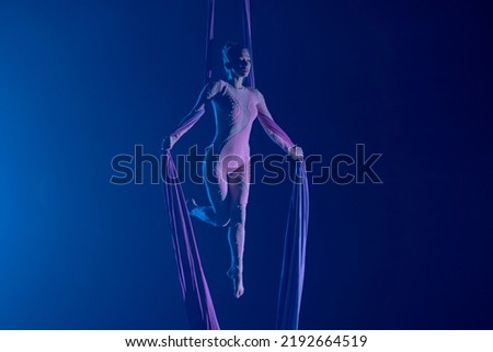 A young woman performing in a circus on aerial silk in the dark with blue light. A female equilibrist balancing on a height. Performance of aerial gymnast. Royalty-Free Stock Photo #2192664519