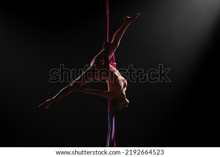Female circus gymnast hanging upside down on aerial silk and demonstrates stretching. Woman performs tricks at height on red silk fabric. Difficult acrobatic stunts on black background with Royalty-Free Stock Photo #2192664523