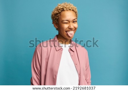 Portrait of funny black girl with blond curly hair showing tongue winking with one eye, making faces, fooling around, aping with silly face, joking, teasing, isolated on blue studio background Royalty-Free Stock Photo #2192681037
