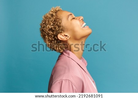 Profile view of black millennial girl laughing out loud on blue studio background dressed in pink shirt, having blond short haircut and wearing stylish nose ring. Sincere positive emotions Royalty-Free Stock Photo #2192681091