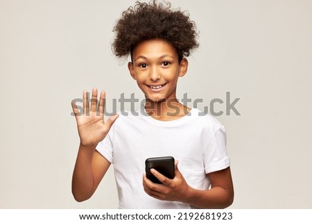 Studio image of happy excited afro boy of 12 waving hand greeting you holding smartphone on gray studio background, video chatting with his friends, using learning application to study online