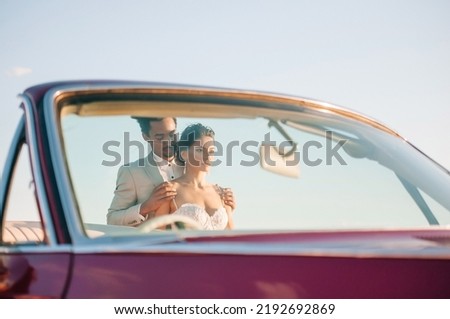 Young couple in love posing at sunset in nature on their wedding day near a red car