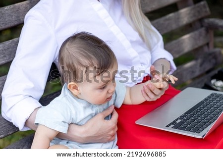 young mother using laptop and sitting with little baby on park bench,outside.freelancer job during maternity concept.business lady working,typing on keyboard.baby make different faces multitasking mom