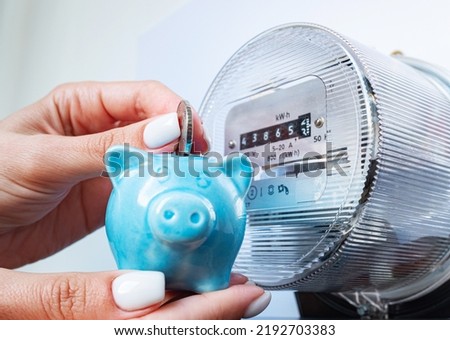 Coin and piggy bank in woman's hands near electricity meter at home. Symbolic image of energy costs and electricity prices, saving the household budget on paying utility bills. Royalty-Free Stock Photo #2192703383