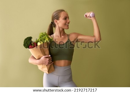 Sporty beautiful woman holding a bag of vegetables and a dumbbell on a green background. The concept of sports and healthy eating.