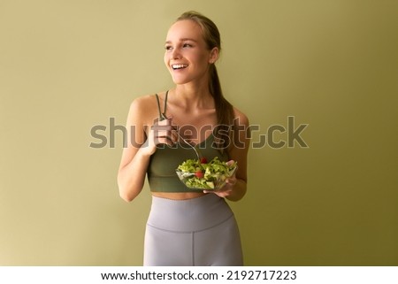A young sporty woman wearing a green top and gray leggings is holding a plate of salad in her hands, looking to the side and smiling while standing near a green background. Royalty-Free Stock Photo #2192717223