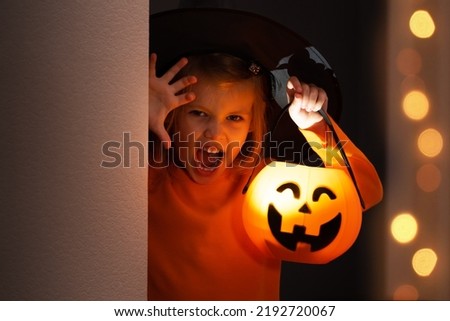 Halloween. Little girl in witch costume hides and scares from corner with lantern pumpkin. Child in black hat plays in horror stories in dark
