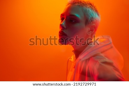 Young androgynous woman in futuristic raincoat with short hair looking away while standing under bright neon light against orange background Royalty-Free Stock Photo #2192729971