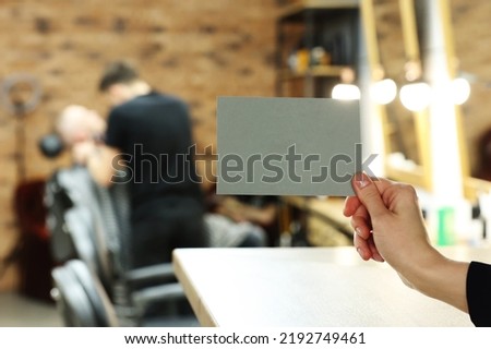 female hand holding grey postcard or bossiness card with mock up on stylish barbershop or beauty salon background. blurred barber man is cutting and shaving the beard o client