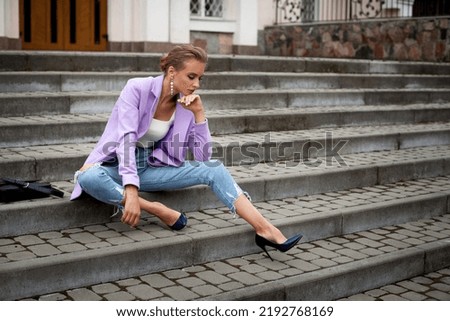 A stylish blonde girl in a white tank top, purple jacket and blue jeans, with long legs, sits on the stairs in the city. There is a leather bag next to it.