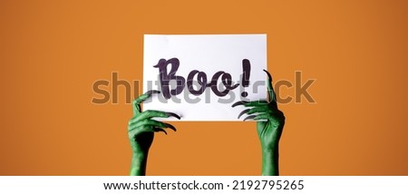 Hands of witch holding paper with text BOO on orange background. Halloween celebration
