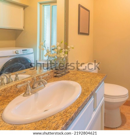 Square Small laundry and bathroom interior with narrow window. There are two laundry units and wall cabinets on the left across the vanity sink beside the toilet with picture frame on the right.
