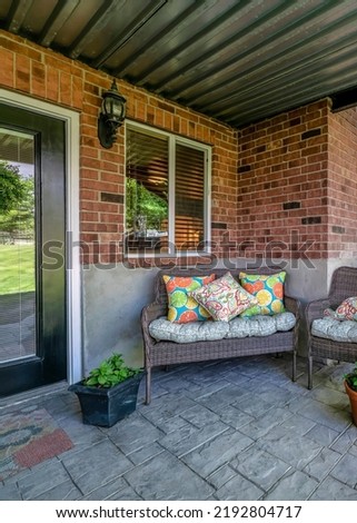 Vertical Woven couch and armchairs on an outdoor patio under the deck of a house with bricks. There is a glass front door on the right side near the windows and a view of a stairs on the left.