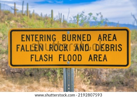 Signage with Entering Burned Area Falling Rocks and Debris Flashflood Area at Tucson, AZ. Close-up of a yellow sign post against the blurred image background of wild plants.