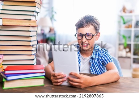 Portrait of a smart schoolboy with a tablet computer. A boy child is sitting at a table with stacks of books, doing homework, using a digital tablet for online lessons and searching for information.