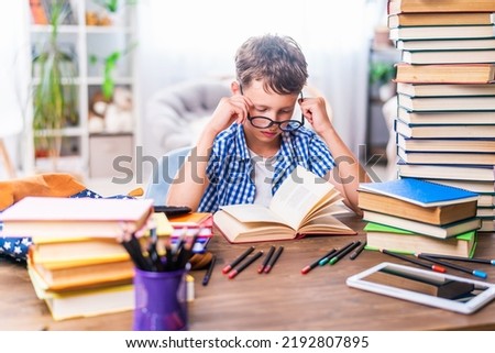 schoolboy boy sees poorly, tries to read through glasses. The child is sitting at home at his desk and reading a book. Back to school. Problems with the eyes. Poor eyesight Royalty-Free Stock Photo #2192807895