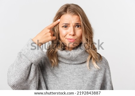 Close-up of confused blond girl in grey sweater, pointing finger at head, scolding something for acting stupid, standing over white background