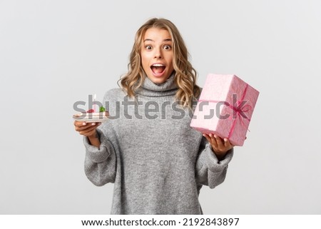 Image of attractive blond girl celebrating birthday, looking surprised, holding b-day cake and a gift, standing over white background