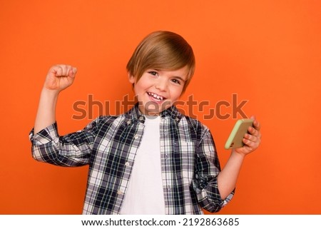 Photo of young cheerful boy rejoice victory champion win competition fists hand use mobile isolated over orange color background