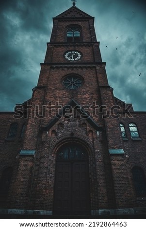 in the photo there is a brick church with a cross on the tower. it is cloudy and the sky is completely covered with clouds. Birds fly over the church. The church has large doors, windows and clock.
