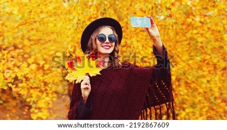 Autumn portrait of stylish happy smiling young woman taking selfie with smartphone and yellow maple leaves wearing round hat, brown knitted poncho in the park