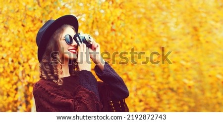 Autumn portrait of happy smiling young woman photographer with film camera wearing round hat, knitted poncho in the park on yellow leaves background