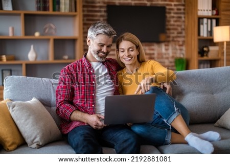 Cheerful mature caucasian wife show to man to laptop, watching video together, sit on sofa, in living room interior. Sale for shopaholic at home, ad and internet offer, video call and modern gadget Royalty-Free Stock Photo #2192881081