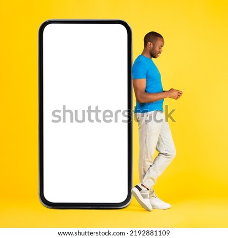 African American Man Using Mobile Phone Posing Near Big Smartphone With Empty Screen Over Yellow Studio Background. Great Application Concept. Square Shot, Full Length