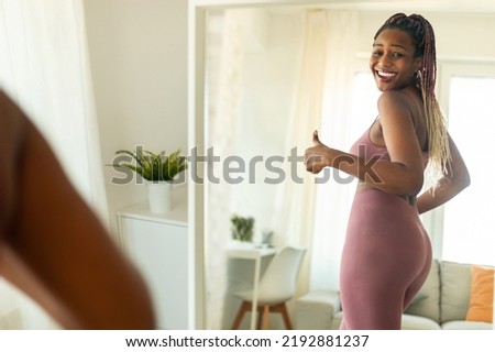 Excited black lady standing in front of mirror after training, happy with result of her diet and exercises, showing thumb up and smiling. Emotional lady achieving weight loss goal, success Royalty-Free Stock Photo #2192881237