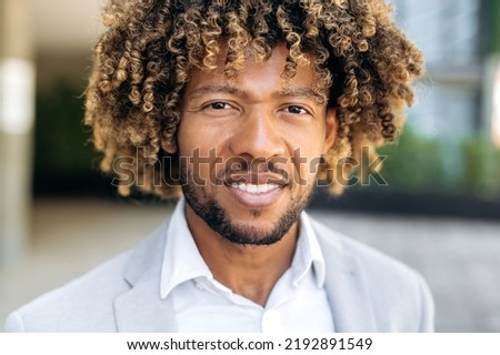 Close-up photo of a handsome charismatic confident curly haired brazilian or hispanic man, successful entrepreneur, standing outdoors, looking at the camera, smiling friendly
