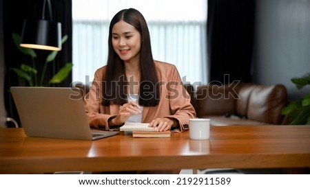 Charming young Asian businesswoman or female office employee working in her office, using laptop computer to research some information on internet.