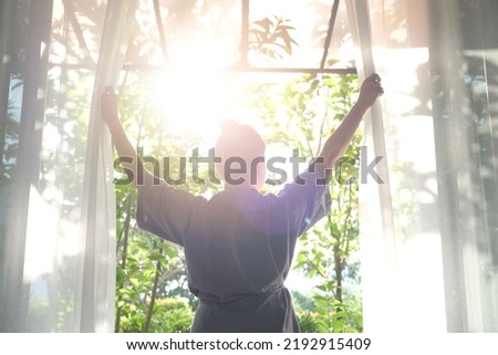 The woman was at the window in the bedroom. She opened the curtains on the window. In the morning and she looks at the view of mountains and trees at sunrise. Royalty-Free Stock Photo #2192915409