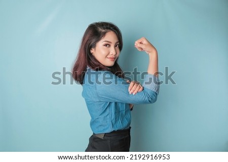 Excited Asian woman wearing a blue shirt showing strong gesture by lifting her arms and muscles smiling proudly Royalty-Free Stock Photo #2192916953