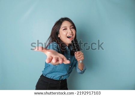 Portrait of carefree Asian woman, having fun karaoke, singing in microphone while standing over blue background Royalty-Free Stock Photo #2192917103