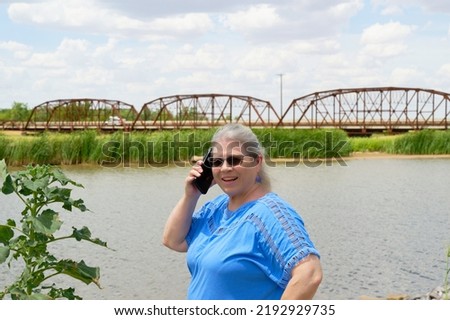 Mature, elderly caucasian woman smiling, using a cellphone with a river and bridge in the background.