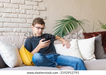 teenage boy in glasses, a gray T-shirt and jeans sits in a relaxed position on the couch with a smartphone in his hands. Communicate with the use of wireless technologies. Online meetings with peers