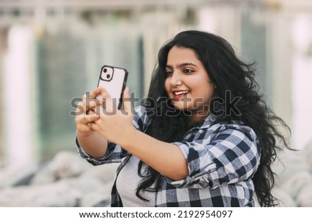 An Indian girl takes a selfie on her phone while sitting on the embankment while walking in the city.