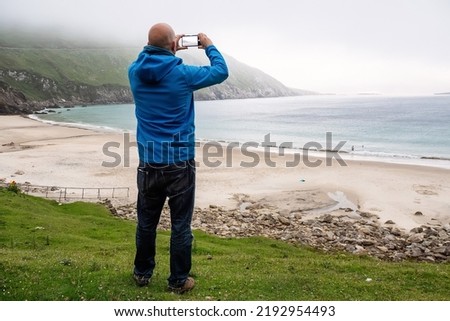 Male tourist taking picture on phone of stunning nature scenery with sandy beach, ocean and mountain. Low cloud. Keem bay, county Mayo, Ireland. Travel concept. Irish landscape.