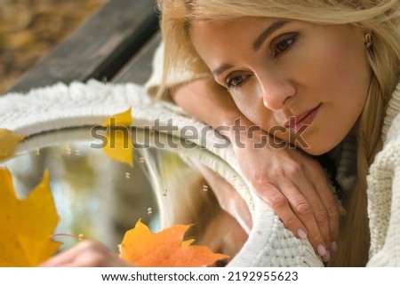 Attractive blonde woman leaned  against the mirror with autumn leaves,reflection of her face.Autumn concept.Beauty in nature.Slow living.Female aging.