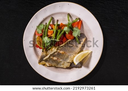 Fried fish with pods, lemon, parsley, tomato and mushroom on a white plate on a black stone background