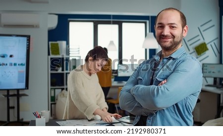 Portrait of confident man entering frame posing with arms crossed and smiling in busy startup office. Relaxed employee acting casual looking at camera in marketing department of small company.