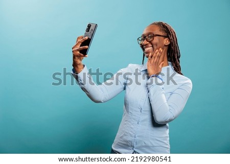 Happy confident african american woman with modern touchscreen smartphone taking selfie photo. Excited pleased young adul person smiling at phone camera while taking picture for social media.
