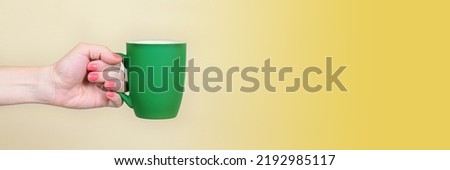 Woman's hand holds a green mug with a matte surface on a neutral beige background. Template for inserting text or images on the mug. Good morning concept. Banner with free space for text.