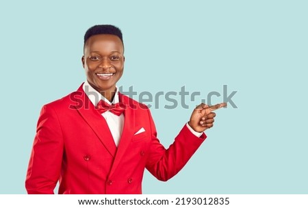 Advertise here. Joyful stylish african man presenting new product pointing finger at copy space on pastel light blue background. Smiling man in red suit with bow tie advertises on banner background. Royalty-Free Stock Photo #2193012835