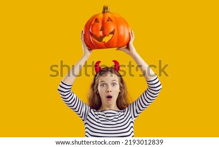 Studio shot of funny girl with Halloween pumpkin. Young woman in devil horns isolated on yellow background, holding up orange pumpkin and looking at camera with funny silly surprised face expression