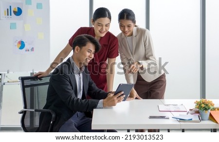 Young asian businessman in suit holding tablet computer while both of his colleague interested in the information on screen. Graph, chart and note are on the presentation board in the background.