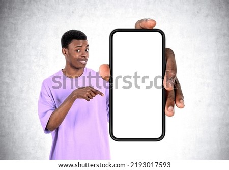 Black young man smiling, finger pointing at large smartphone mock up copy space screen, grey concrete wall background. Concept of connection and social media
